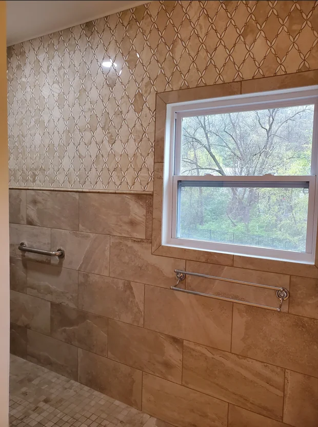 Bathroom shower with travertine tile walls, geometric wallpaper, frameless glass enclosure, and chrome fixtures