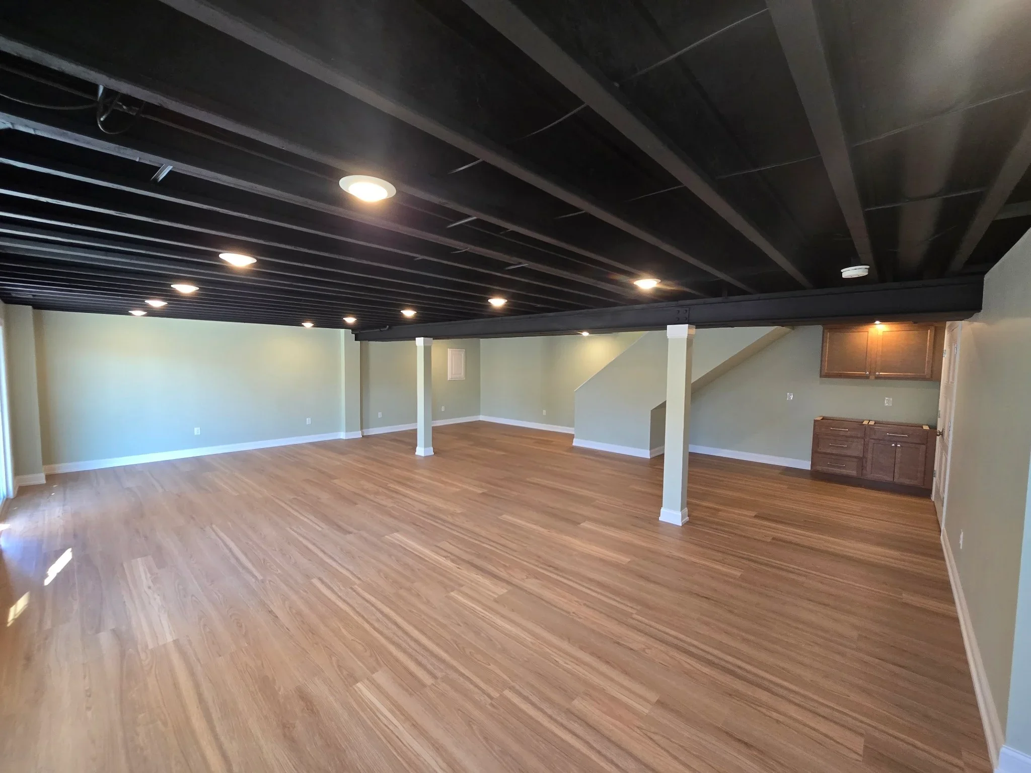 Finished basement with vinyl plank flooring, black drop ceiling, recessed lights, and white support columns